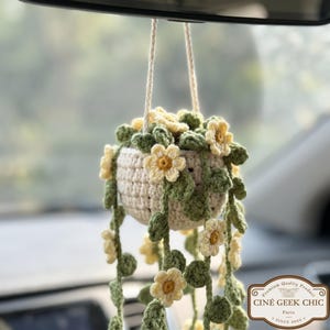Op de afbeelding: Een gehaakte hangende plantenbak met een crèmekleurige pot, versierd met gele bloemen en groene bladeren. De plant hangt aan een crèmekleurig touwtje, wat een charmante, handgemaakte uitstraling creëert. De plantenbak hangt aan de achteruitkijkspiegel van een auto.