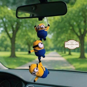 May include: A car charm featuring three crocheted Minion figures, hanging from a car's rearview mirror. The Minions are yellow and blue. The background shows a tree-lined road. The charm is handmade.
