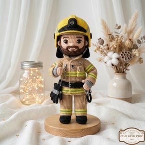 May include: Crocheted firefighter doll wearing a yellow helmet, tan uniform with reflective stripes, and black gloves. The doll has a brown beard and is standing on a wooden base. The doll is holding a key ring and a pair of gloves.