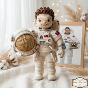 May include: A crocheted astronaut doll in a cream-colored spacesuit, holding a matching helmet. The doll has brown hair and blue eyes. A framed photo of a man in a spacesuit is in the background. The doll has a patch with the Slovakian flag.