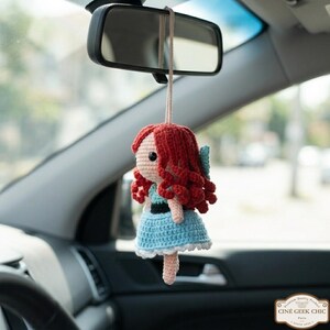 A crocheted doll with red hair and a blue dress is hanging from a car's rearview mirror.