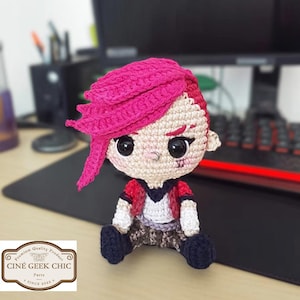 Op de afbeelding: Gehaakte pop van een personage met roze haar, een rode jas en een donkerblauwe broek. De pop heeft een kleine zilveren neusring en een gestikt litteken op het gezicht. De pop zit op een zwart oppervlak. De pop is een product van Cine Geek Chic.
