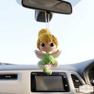 Puede incluir: Un adorno de coche de hada hecho a ganchillo. El hada tiene un moño amarillo, ojos negros grandes y un vestido verde. El adorno cuelga del espejo retrovisor. El texto "Ciné Geek Chic Paris" es visible.
