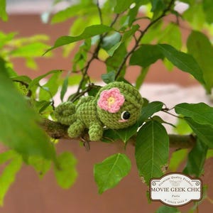 May include: A green crocheted frog with a pink flower on its head. The frog is sitting on a branch of a tree. The frog is a handmade toy.