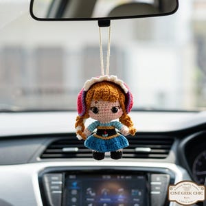 May include: A crocheted doll of a character with orange hair in braids, wearing a blue dress and a red and white hat, hanging from a string. The doll is suspended from a car's rearview mirror.