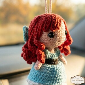 A crocheted doll with red hair and a blue dress is hanging on a string.