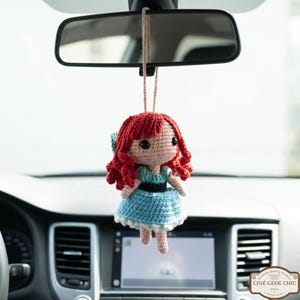 A crocheted doll with red hair and a blue dress is hanging from the rearview mirror of a car.