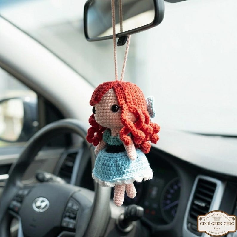 A crocheted doll with red hair and a blue dress is hanging from a car's rearview mirror.