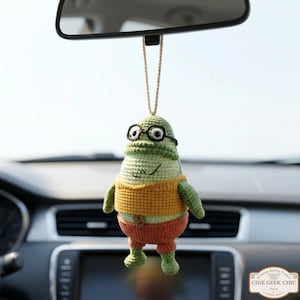 May include: A handmade, green and yellow crochet car air freshener shaped like a cartoon character wearing glasses. The character has orange shorts and a yellow shirt. The air freshener hangs from a string attached to a car's rearview mirror.