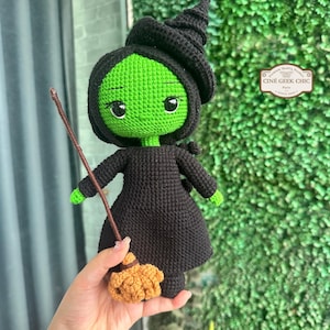 May include: A handmade crochet doll of a green-skinned witch wearing a black dress and hat. The doll has large, round eyes and is holding a small broom. The doll is a whimsical representation of a witch.