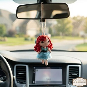 A crocheted doll with red hair and a blue dress is hanging from the rearview mirror of a car.