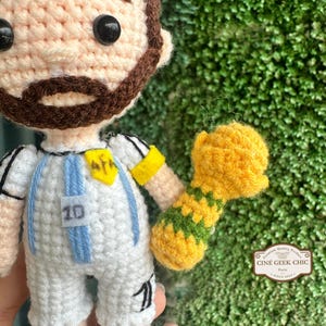 May include: Crocheted doll of a soccer player wearing a white and light blue uniform with the number 10. The doll has a brown beard and hair, and is holding a yellow and green trophy. The doll is approximately 5 inches tall.