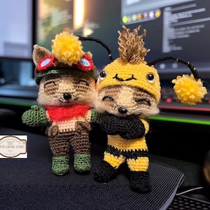 May include: Two handmade crochet figures. One is dressed in a brown outfit with a red scarf and a hat with goggles and a yellow pom-pom. The other is dressed as a bee in yellow and black with a yellow hat and pom-pom. The figures are on a dark surface.