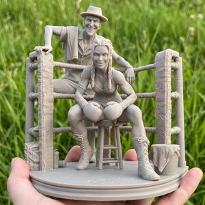 May include: A detailed sculpture depicting a boxing ring. A female boxer, wearing gloves and boots, sits on a stool, with a man in a hat leaning on the ropes behind her. The sculpture is a neutral colour, set against a green background.