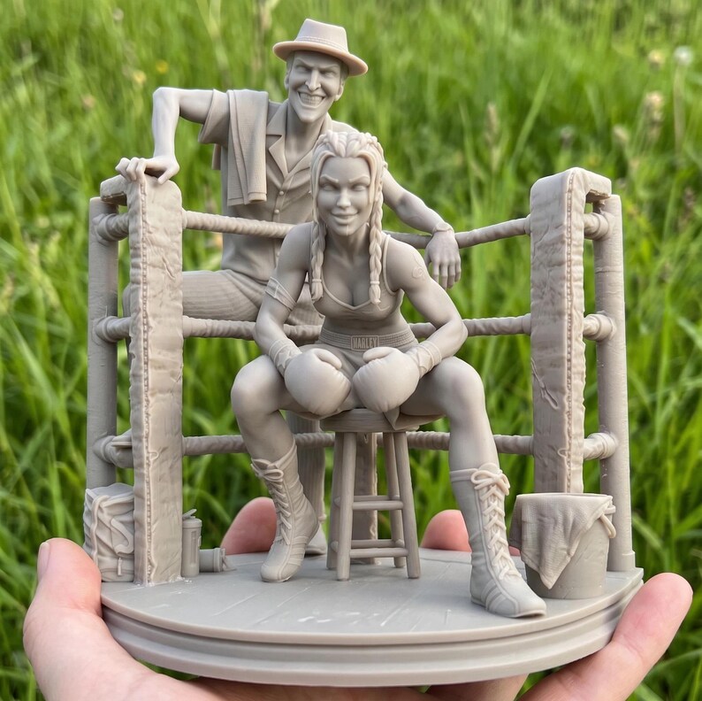 May include: A detailed sculpture depicting a boxing ring. A female boxer, wearing gloves and boots, sits on a stool, with a man in a hat leaning on the ropes behind her. The sculpture is a neutral colour, set against a green background.