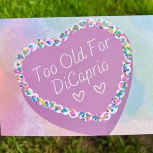 May include: A purple heart-shaped card with a white border and colorful glitter around the edge. The card says "Too Old For DiCaprio" with two white hearts.