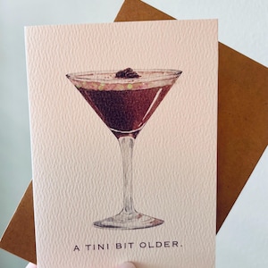 May include: A greeting card with a watercolor illustration of a martini glass filled with a dark brown liquid and a garnish. The card is light pink with the text "A TINI BIT OLDER." A brown envelope is behind the card.