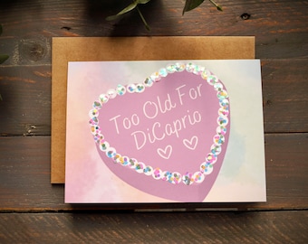 May include: A pink heart-shaped card with glitter around the edges. The card says "Too Old For DiCaprio" with two small hearts below the text.