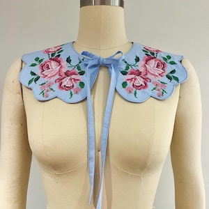 May include: A light blue cotton collar with a scalloped edge and embroidered pink roses. The collar has two long ties that can be tied at the neck.