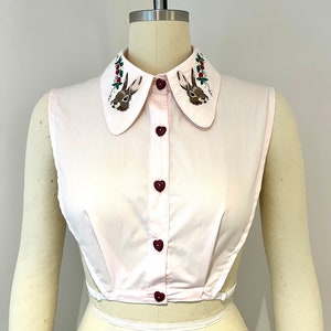 May include: A pink sleeveless blouse with a white collar featuring embroidered bunnies and red berries. The blouse has a button-up front with heart-shaped buttons.
