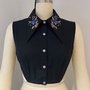 May include: A black sleeveless shirt with a large collar featuring embroidered swallows and flowers. The shirt has a button-up front with lavender buttons.