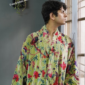 May include: A green velvet robe with a floral and bird print. The robe has a shawl collar and a tie belt. The robe is worn by a person standing near a window.