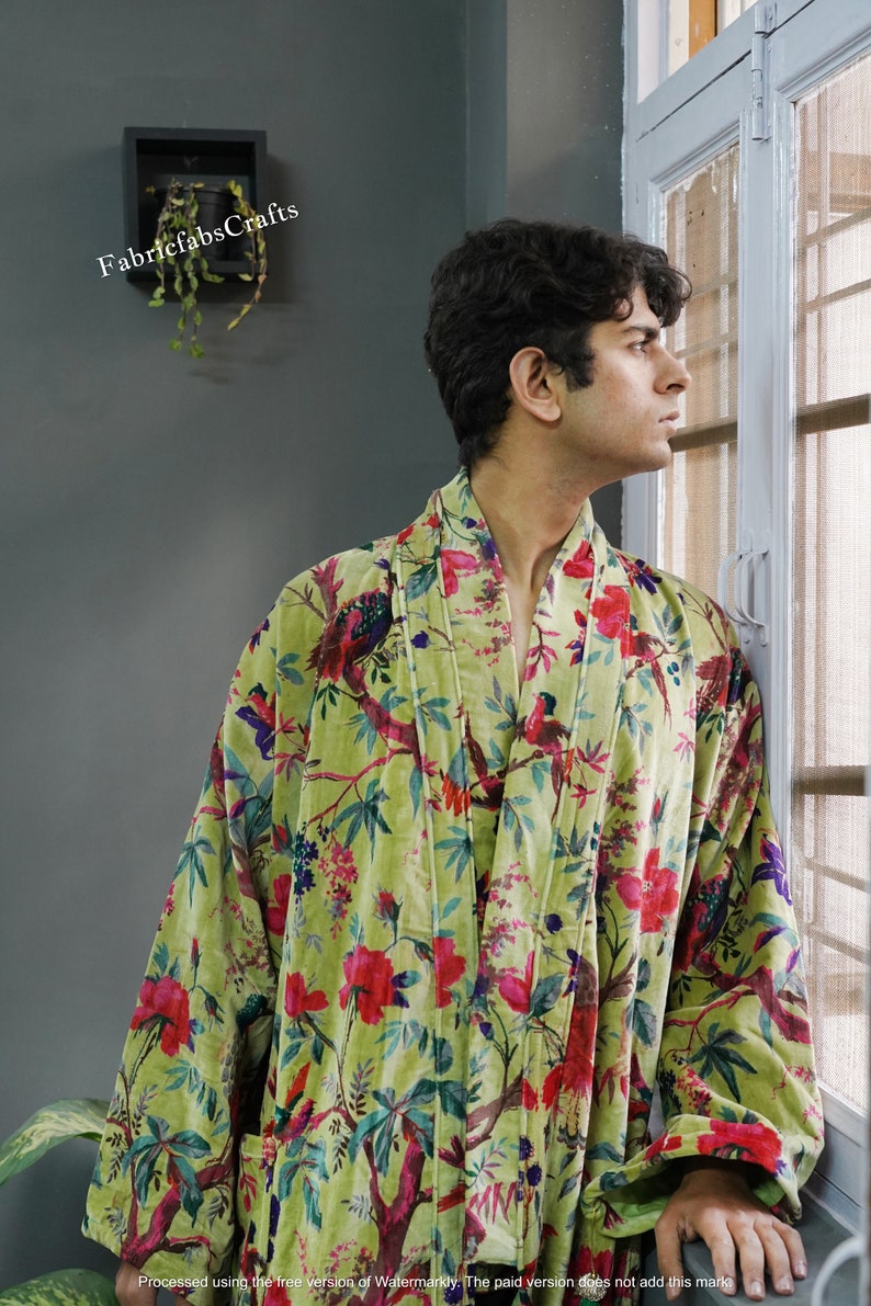 May include: A green velvet robe with a floral and bird print. The robe has a shawl collar and a tie belt. The robe is worn by a person standing near a window.