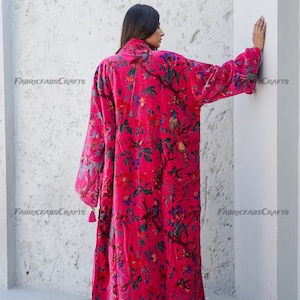 May include: A long, pink velvet robe with a floral print featuring birds and branches. The robe has long sleeves and a tassel detail at the cuffs.