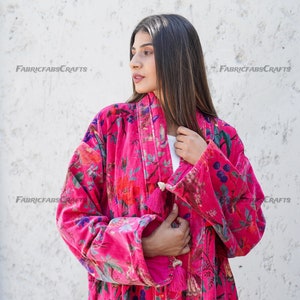 May include: A pink velvet robe with a floral print and tassel details. The robe has long sleeves and a tie closure. The robe is worn by a person with long dark hair.