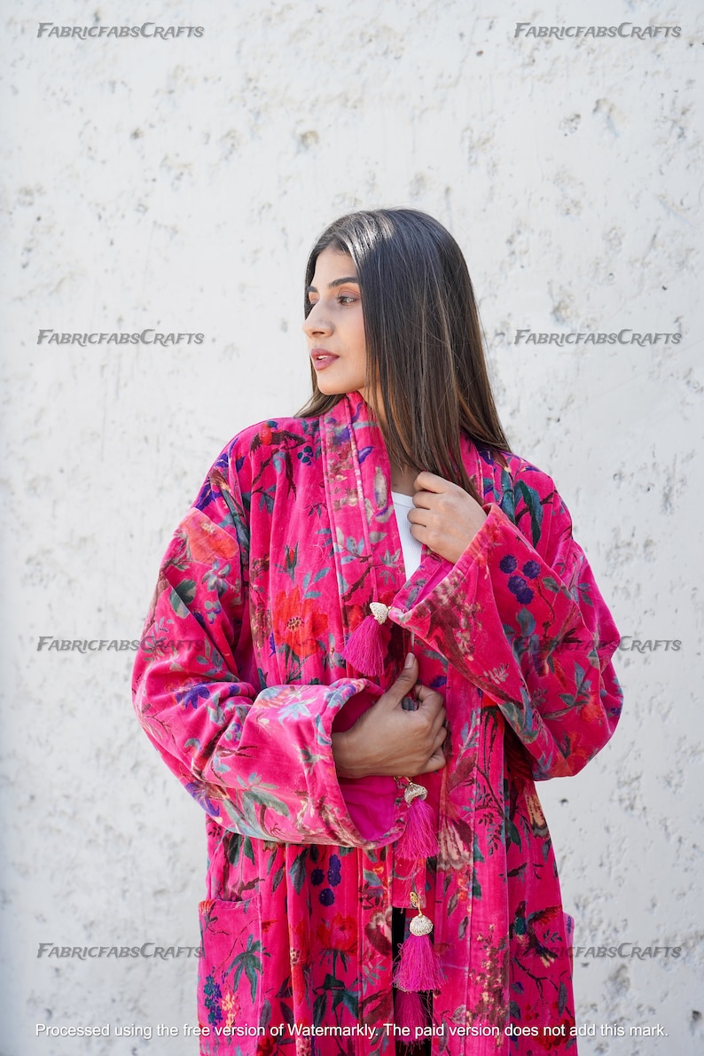 May include: A pink velvet robe with a floral print and tassel details. The robe has long sleeves and a tie closure. The robe is worn by a person with long dark hair.