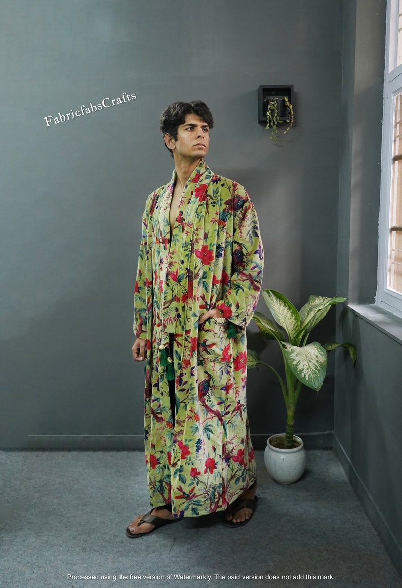 May include: A green velvet robe with a floral print featuring birds and flowers. The robe has a long, flowing silhouette and a sash tie at the waist.