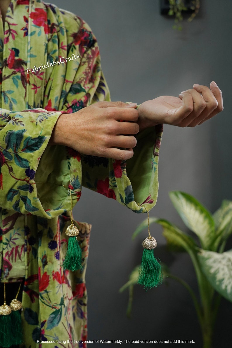 May include: A green velvet robe with a floral print and green tassel accents. The robe is being held by a person's hands.