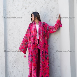 May include: A long, pink velvet robe with a floral print. The robe has a shawl collar, long sleeves, and a tie belt. The robe is worn by a person standing in front of a white wall.