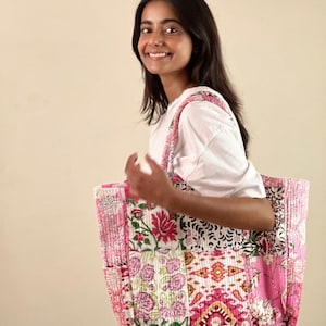 Può includere: Una borsa tote in patchwork rosa e bianco con motivi floreali e geometrici. La borsa ha un ampio scomparto principale e una tasca più piccola sul davanti.