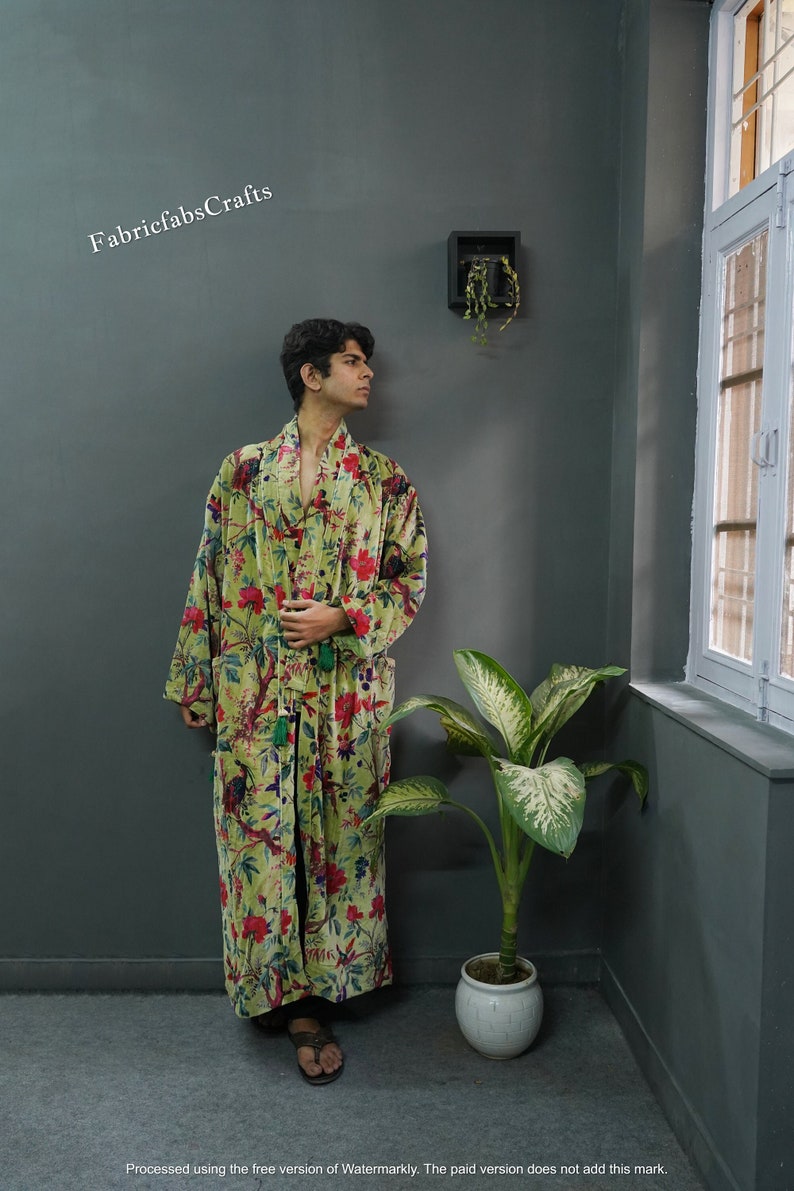 May include: A green velvet robe with a floral print and tassel ties. The robe is worn by a person standing in a room with a plant and a small framed picture on the wall.