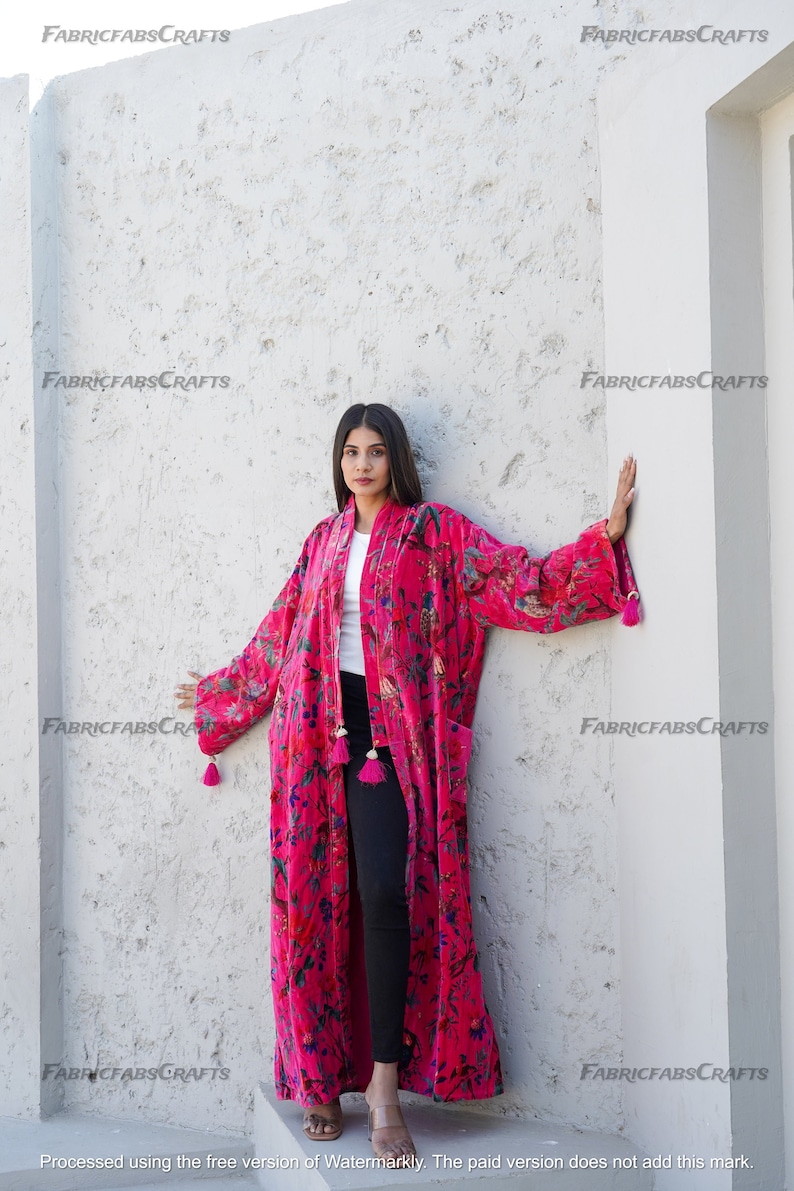 May include: A long, pink velvet robe with a floral print and tassels. The robe is open at the front and the person wearing it is standing against a white wall.