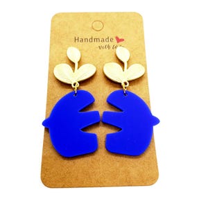 Op de afbeelding: Een paar blauwe vogelvormige oorbellen met goudkleurige bladeren. De oorbellen worden gepresenteerd op een bruine kaart met de tekst "Handmade with love". Een uniek accessoire.