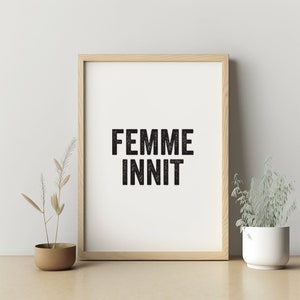 May include: A framed print with the text "FEMME INNIT" in black lettering on a white background. The frame is made of light wood and is sitting on a light wood surface.