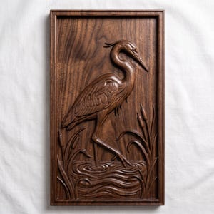 May include: A rectangular dark brown wooden wall plaque featuring a detailed relief carving of a heron standing in water among reeds. The wood grain is visible throughout the piece, which is framed by a simple, raised border edge.