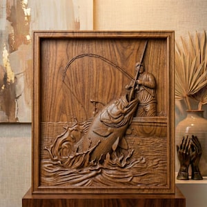 May include: A wooden relief carving depicting a fisherman in a boat reeling in a large fish. The artwork is framed and features detailed carving of water, fish, and the fisherman. The wood has a warm, natural tone, and the scene is set within a square frame.