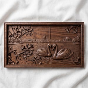 May include: A rectangular wooden relief carving depicting two swans in a pond with the Washington Monument and the Capitol Building in the background. Cherry blossoms frame the scene. The carving is in a dark brown color.