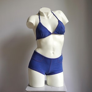 May include: A blue bikini set displayed on a mannequin. The two-piece swimsuit features a triangle top and high-waisted shorts. The edges of the bikini are accented with red stitching. The mannequin is white and has a neutral background.