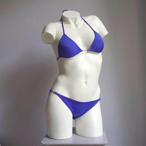 Pode incluir: Um conjunto de biquíni azul royal exibido em um manequim branco. A parte de cima triangular e a parte de baixo de cintura baixa são conectadas com detalhes dourados. O traje de banho é projetado com gola halter e corte de biquíni clássico.