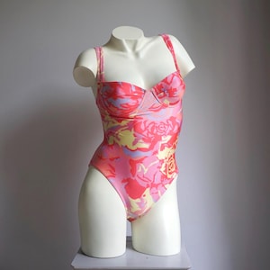 Può includere: Un costume da bagno intero con fantasia floreale nei toni del rosa, giallo e viola. Il costume da bagno ha una scollatura a cuore e spalline sottili. L'indumento è esposto su un manichino bianco.