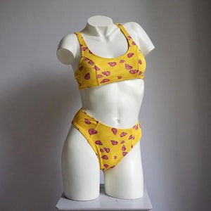 Könnte beinhalten: Gelber zweiteiliger Badeanzug mit einem sich wiederholenden rosa Muschelmuster. Das Oberteil ist im Sport-BH-Stil gehalten, und die Unterteile sind hoch geschnitten. Der Badeanzug wird auf einem weißen Mannequin-Torso vor grauem Hintergrund präsentiert.