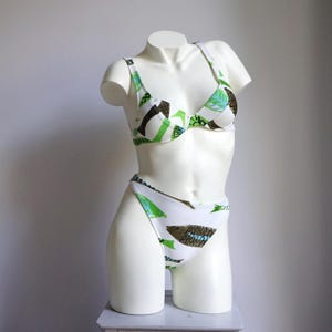 May include: White bikini set with a green, blue, and brown abstract pattern. The two-piece swimsuit features a triangle top and high-waisted bottoms. The bikini is displayed on a white mannequin.