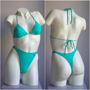 May include: A turquoise two-piece bikini on a mannequin. The triangle top has thin straps, and the high-cut bottoms have a small embroidered detail. The bikini is displayed from the front and back, showing the tie closures.