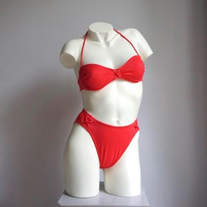 May include: A vibrant red bikini set displayed on a white mannequin. The top is a bandeau style with a halter neck tie, and the bottoms are high-waisted. Both pieces feature decorative lacing details. The mannequin is positioned against a neutral gray background.