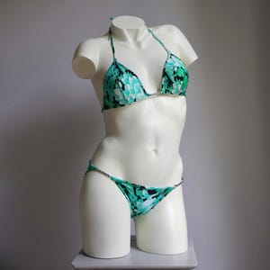 May include: A two-piece bikini set with a floral pattern in shades of teal, black, and white. The bikini top has a halter neck design, and the bottoms are a classic string style. The swimwear is displayed on a white mannequin.