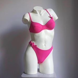 May include: A bright pink bikini set displayed on a white mannequin. The two-piece swimsuit features a classic bra top with adjustable straps and a matching high-cut bottom. Small bow accents adorn the straps and the side of the bottoms.